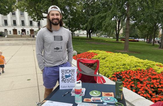 A photo of Dylan Duerre, the Madison ambassador for the Campaign to Save the Boundary Waters, posing in front of the table he operates at the Madison Farmers Market.
