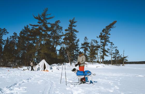 A man and dog play in the snow outside a canvas winter camping tent.