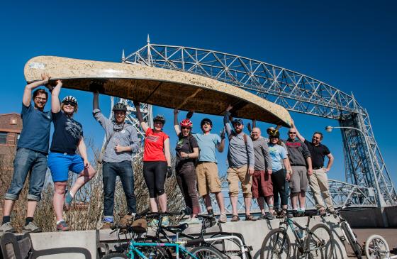 photo of 11 people holding up a tan Wenonah canoe infront of the Duluth Canal Park Bridge