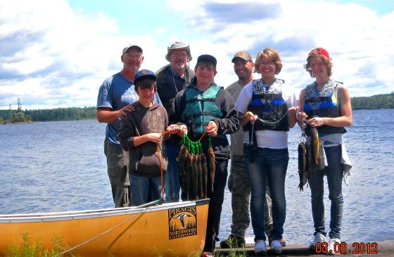 Photo of 7 people standing next to a canoe on the edge of a lake holding fish and smiling