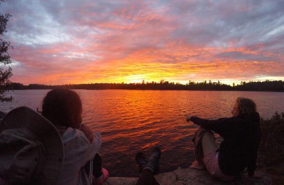 photo of 2 people sitting infront of a bright orange/pink sunset over water