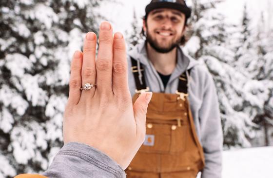 Photo of woman holding left hand out infront of camera with engagement ring on it with man standing behind hand smiling