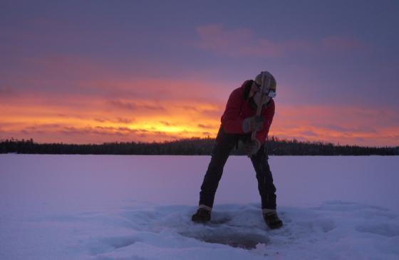 Photo of Amy Freeman digging an ice hole on a frozen lake next to a Boundary Waters sunset