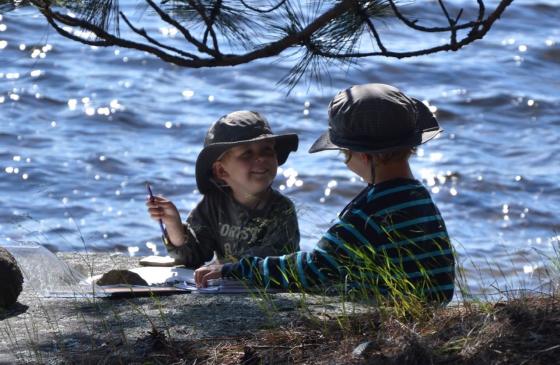 Photo of two children sitting by water in bucket hats under a tree smiling at eachother