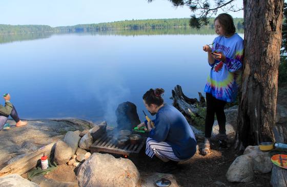 Photo of someone squatting nexto a campfire with someone next to them leaning against a tree with a lake in the background