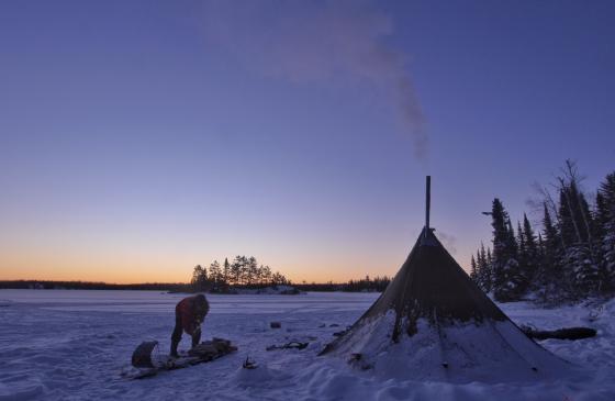 Photo of canvas tent silhouette and person leaning over sled on frozen lake infront of sunset