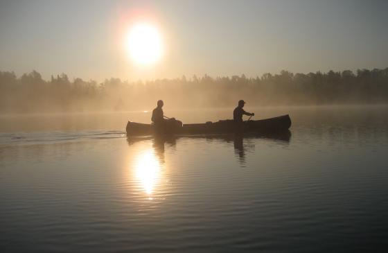 Silhouette of 2 people paddling canoe with sun shining in background