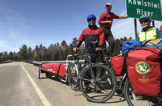 photo of 3 people standing next to 2 bikes with Northstar Canoe packs strapped on them, and a red canoe in the back attached to the bike