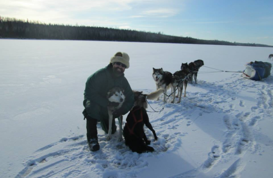 Photo of man kneeling on snow next to sled dogs