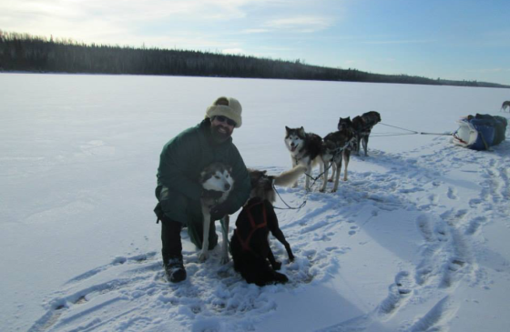 Photo of person kneeling next to a sled dog on a frozen lake with sled dogs in the background