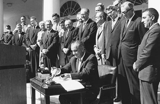 Black and white photo of Lynden B Johnson signing the Wilderness Act of 1964 with a row of men standing behind him in suits 