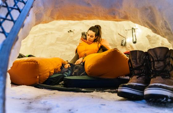 Photo of person sitting in an orange sleeping bag leaning against an igloo from the inside with dog laying next to them and big winter boots in the foreground