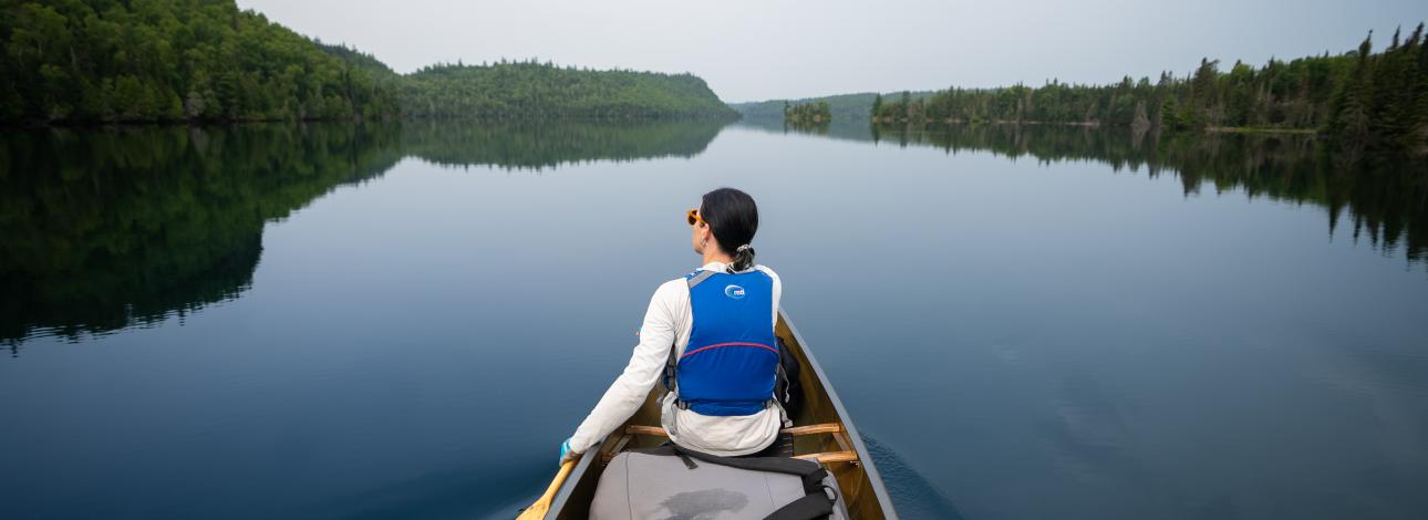 View from behind a woman canoeing on wilderness lake that is a calm and still with palisades in the distance.